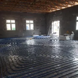 Niedokończony pokój z widoczną instalacją ogrzewania podłogowego z niebieskich rurek na folii, ściany z szarego betonu komórkowego, drewniany strop, okna z białymi ramami.