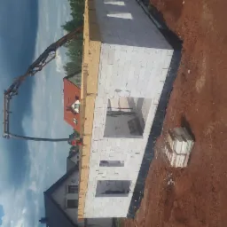 Budowa domu z białych bloczków: widok na wznoszone ściany parteru z użyciem pompy do betonu, w tle zabudowa jednorodzinna i pochmurne niebo.