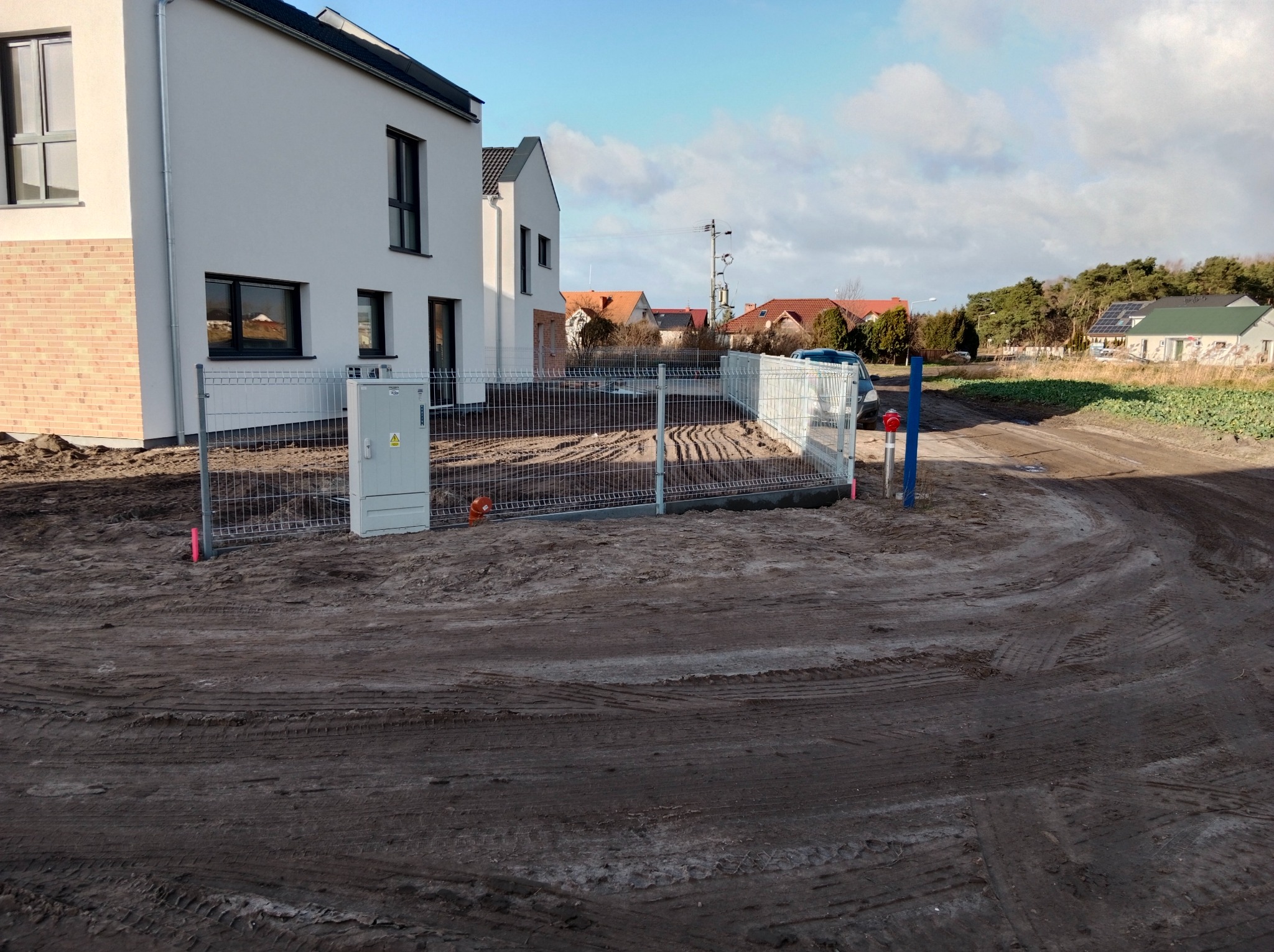 Nowoczesne, ocynkowane ogrodzenie panelowe przed nowo wybudowanym domem jednorodzinnym, widoczne ślady prac ziemnych i tymczasowa skrzynka elektryczna obok słupka.