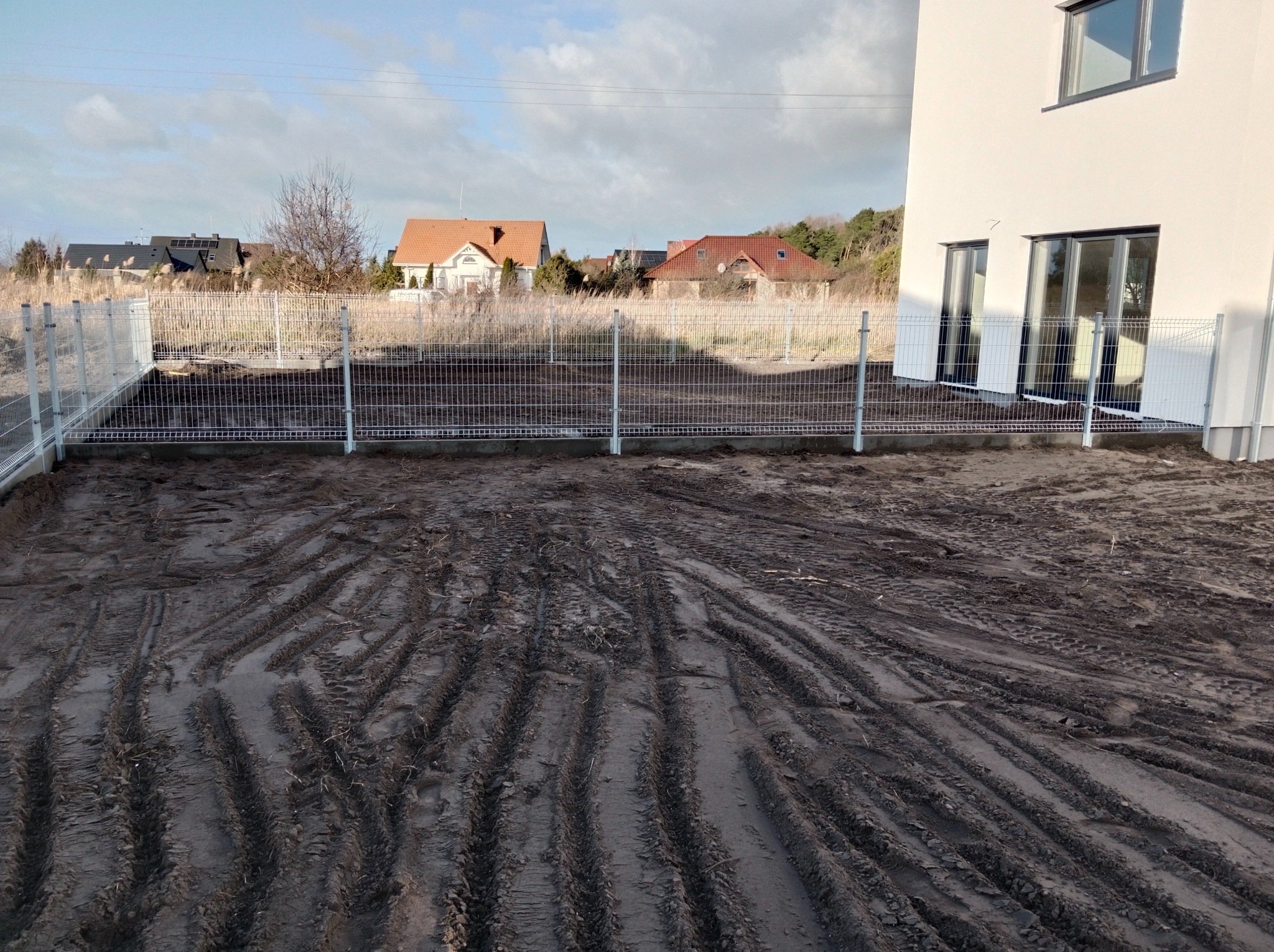 Nowoczesny dom z białymi ścianami i szarymi oknami, otoczony metalowym ogrodzeniem panelowym na jeszcze niezagospodarowanym terenie z widocznymi śladami opon