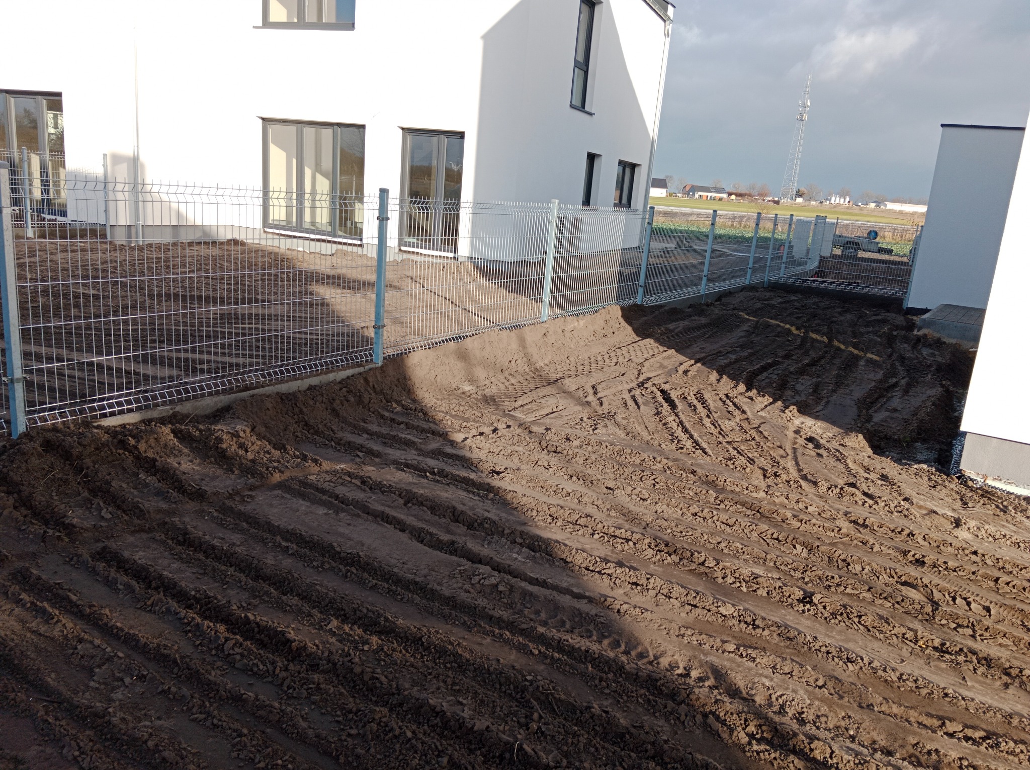 Świeżo zamontowane ogrodzenie panelowe z metalowej siatki otaczające teren wokół nowoczesnego, białego domu. Ziemia przygotowana pod trawnik, widoczne ślady po maszynach rolniczych.