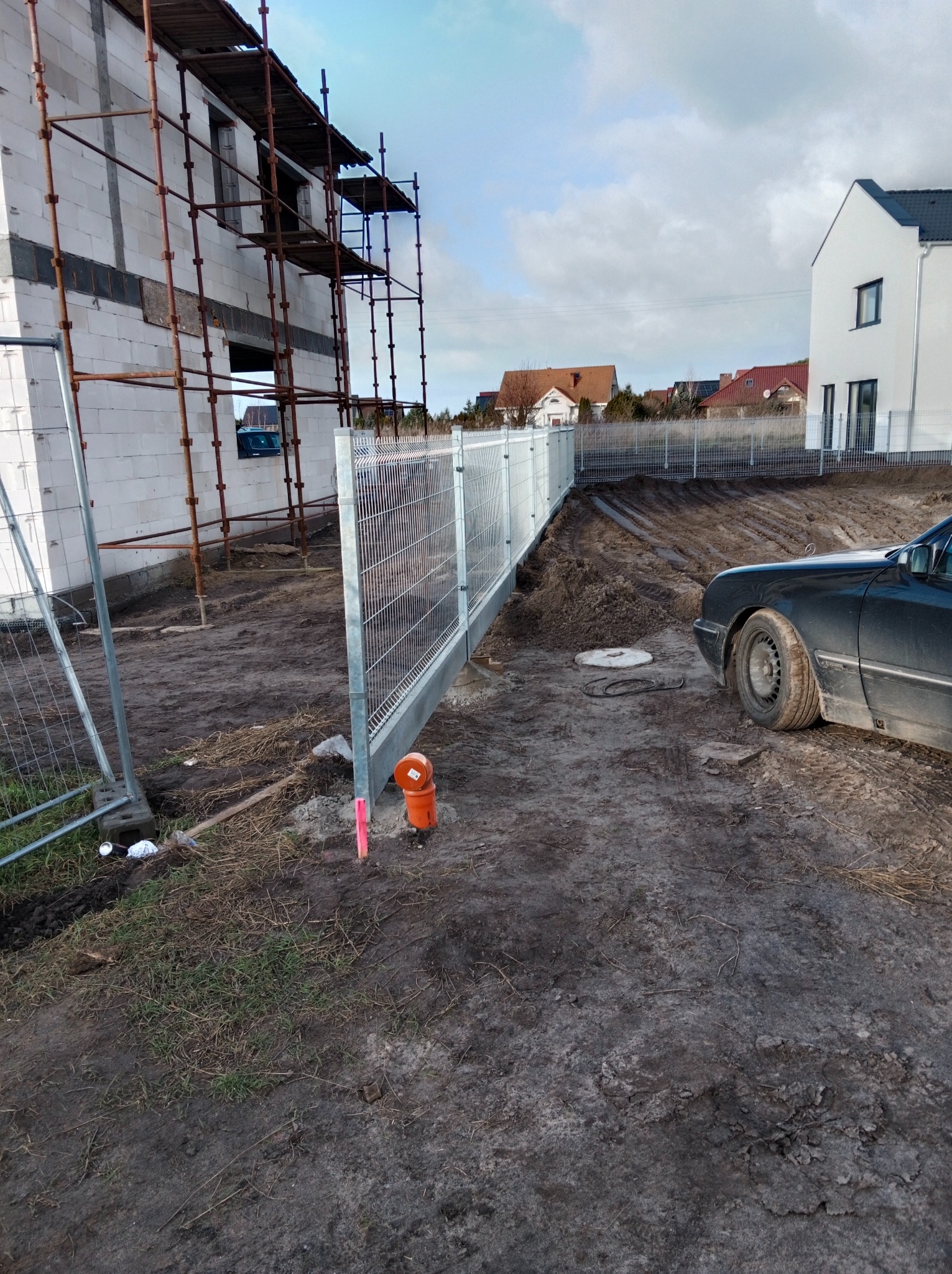 Fragment metalowego ogrodzenia panelowego w trakcie montażu na nierównym terenie budowy, widoczny budynek w trakcie wznoszenia z rusztowaniami oraz zaparkowany samochód osobowy.