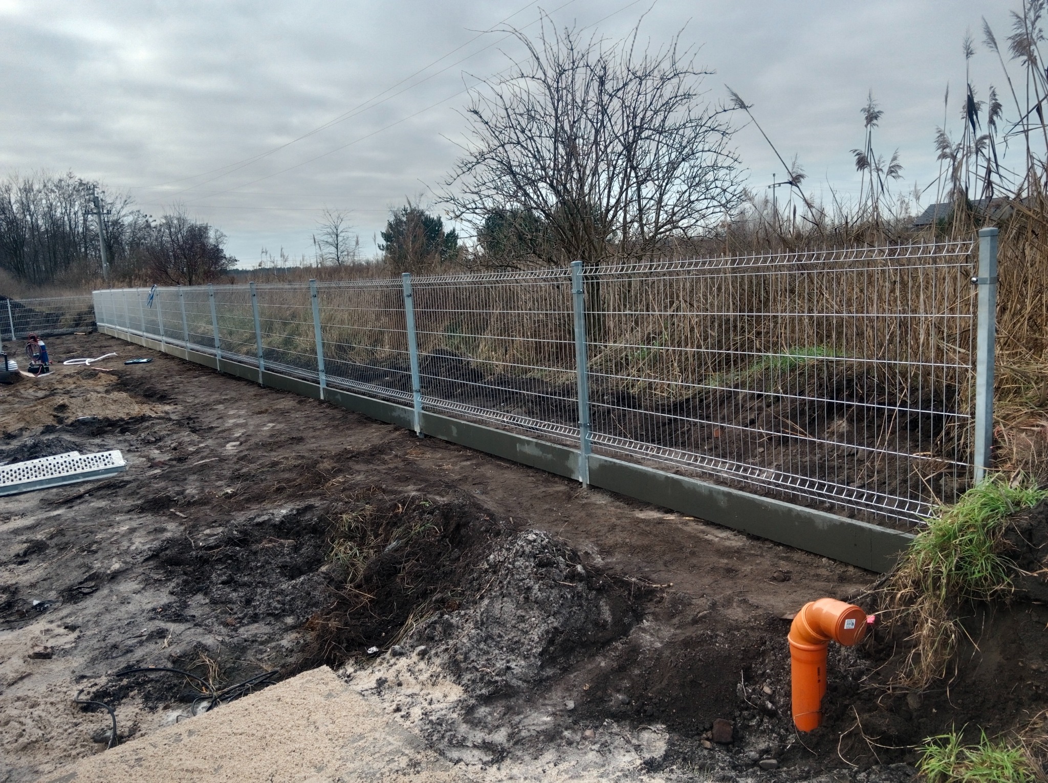 Długi, panelowy, metalowy płot z betonową podmurówką, w trakcie montażu, na tle łąki i drzew, z widoczną rurą PCV w ziemi.
