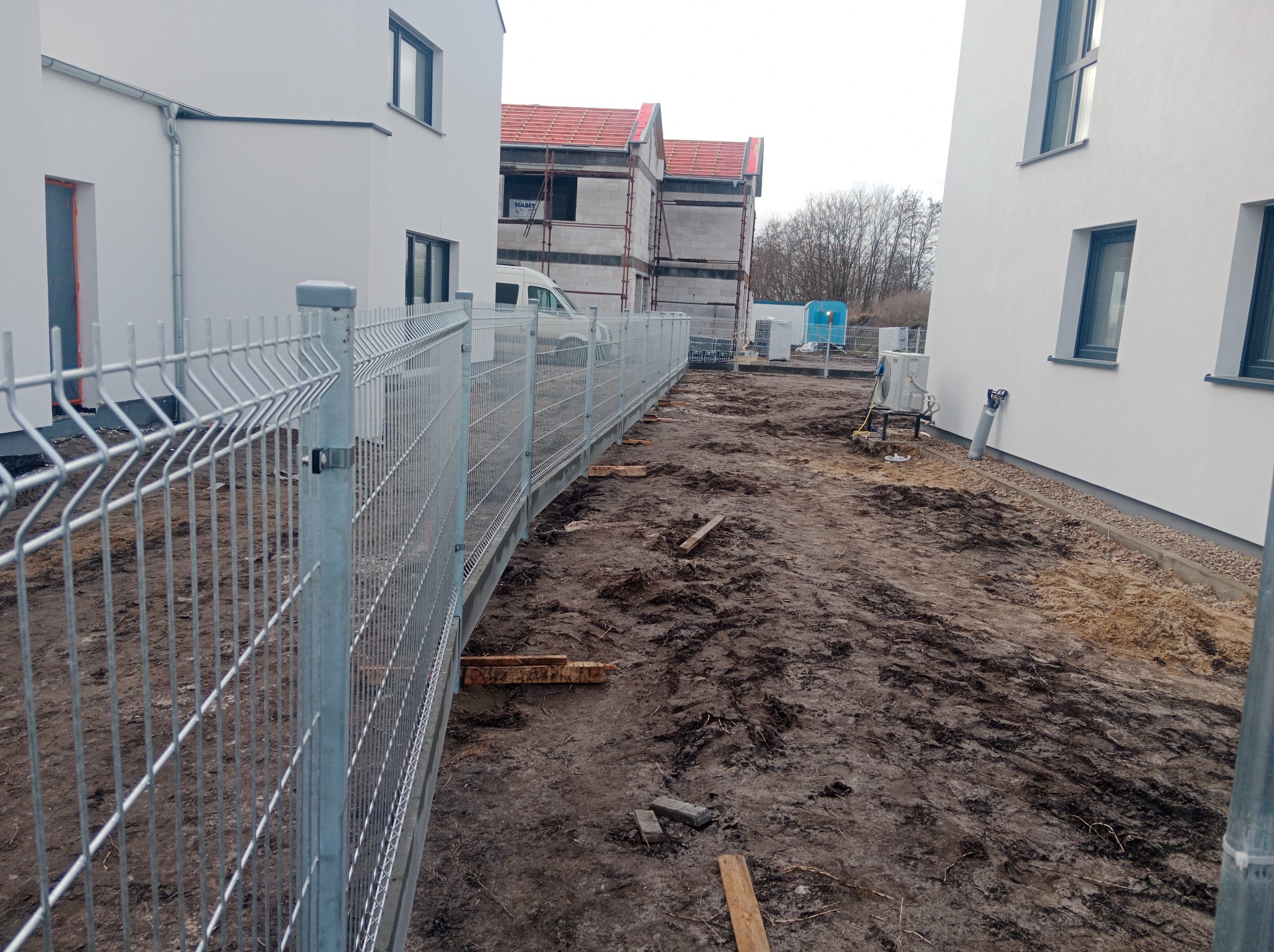 Długi, metalowy, panelowy system ogrodzeniowy wzdłuż błotnistego terenu budowy z nowymi białymi budynkami w tle.