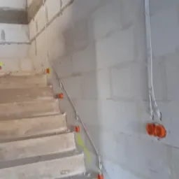 Kable elektryczne w peszlach i pomarańczowe puszki instalacyjne prowadzone wzdłuż surowych schodów z betonu komórkowego, widok z dołu.