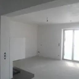 Surowe wnętrze pokoju po nałożeniu gładzi gipsowej na ściany i sufit, widoczne puszki elektryczne, okno balkonowe z widokiem na zewnątrz, szare podłoże.