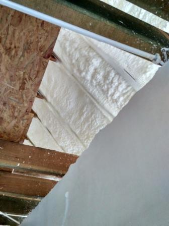 Aplikacja izolacji natryskowej w przestrzeni między krokwiami dachu, widoczne fragmenty drewnianej konstrukcji i biała, spieniona izolacja z nierówną powierzchnią.