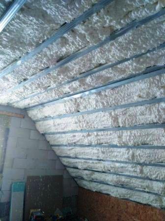 Skośne poddasze z metalową konstrukcją wypełnione białą, nierówną warstwą izolacyjną, widok z dołu.