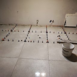 Łukasz - Układanie jasnoszarych płytek podłogowych z użyciem systemu poziomowania, widoczny biały pojemnik i fragment ściany z instalacją elektryczną.