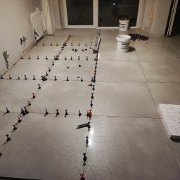 Łukasz - Układanie jasnoszarych płytek podłogowych z użyciem systemu poziomowania, widoczne krzyżyki dystansowe i wiaderka z zaprawą w tle.