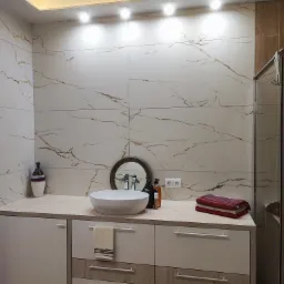 Nowoczesna łazienka z białymi płytkami imitującymi marmur z brązowymi żyłami, okrągła umywalka nablatowa, szafka pod umywalką z białymi frontami i drewnianymi akcentami, oświetlenie punktowe...