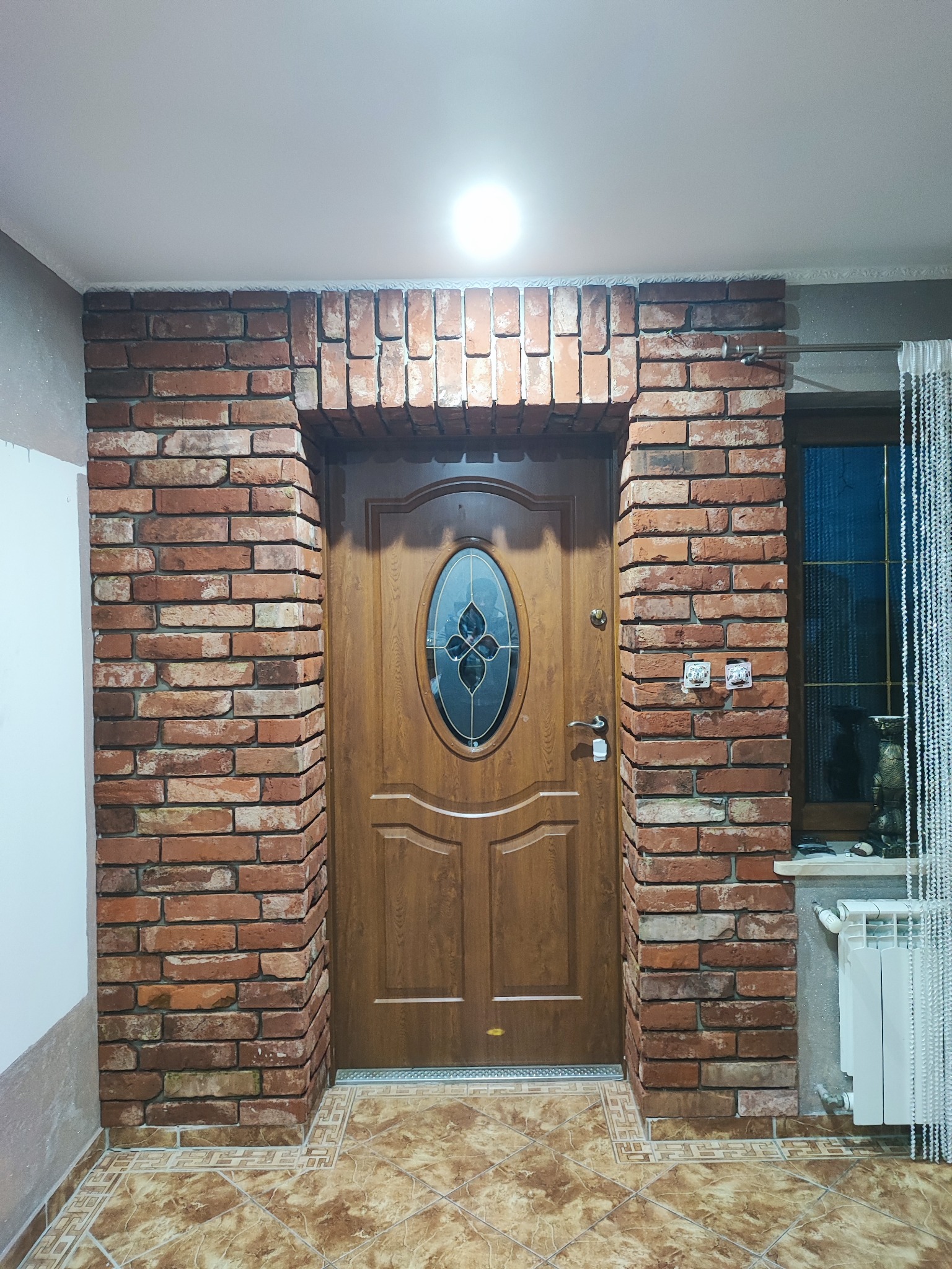 Wejście do pomieszczenia z drewnianymi drzwiami z owalnym przeszkleniem, otoczone dekoracyjną cegłą, z widocznymi elementami wykończenia, takimi jak płytki podłogowe i grzejnik.