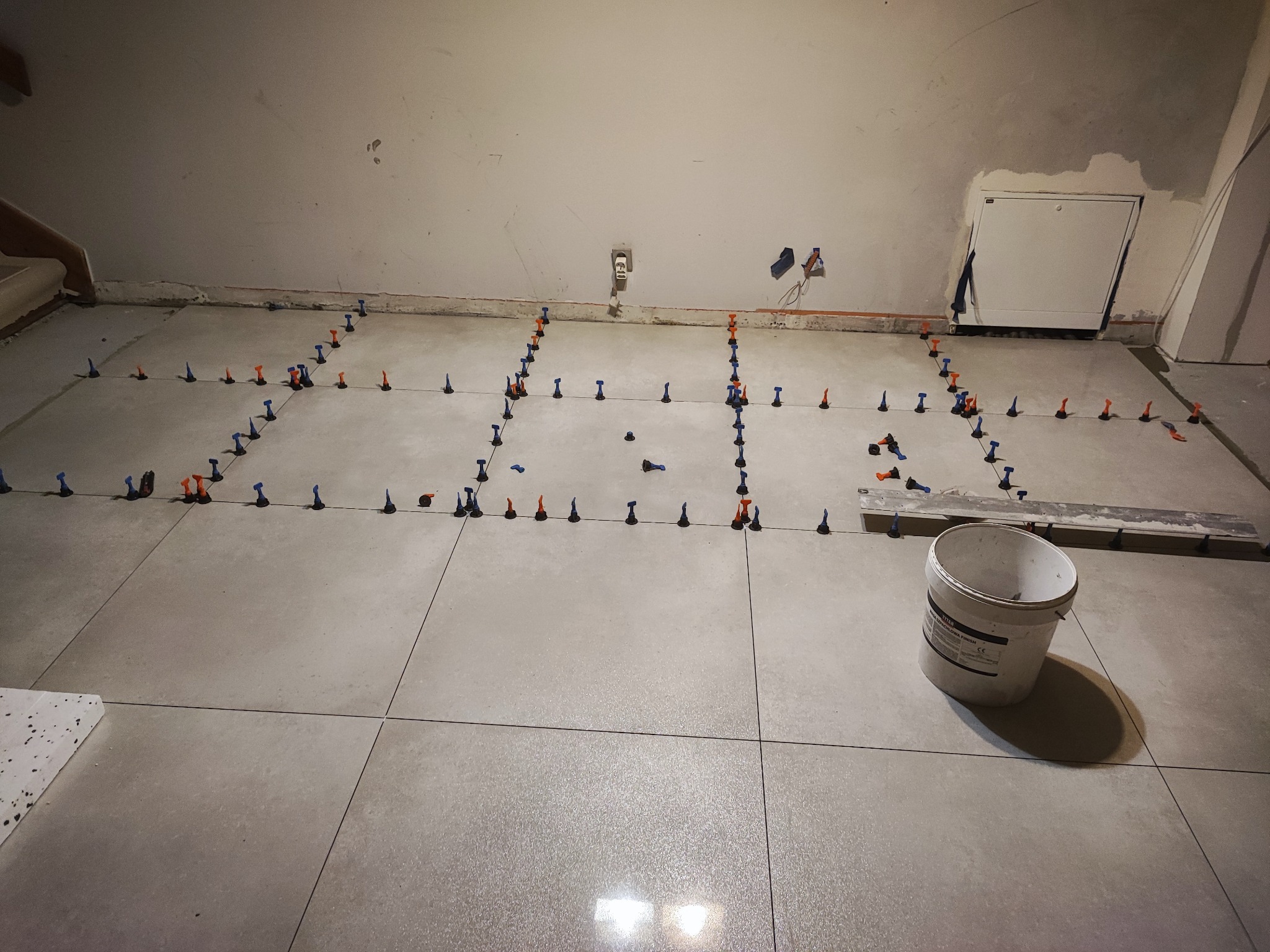 Układanie jasnoszarych płytek podłogowych z użyciem systemu poziomowania, widoczny biały pojemnik i fragment ściany z instalacją elektryczną.