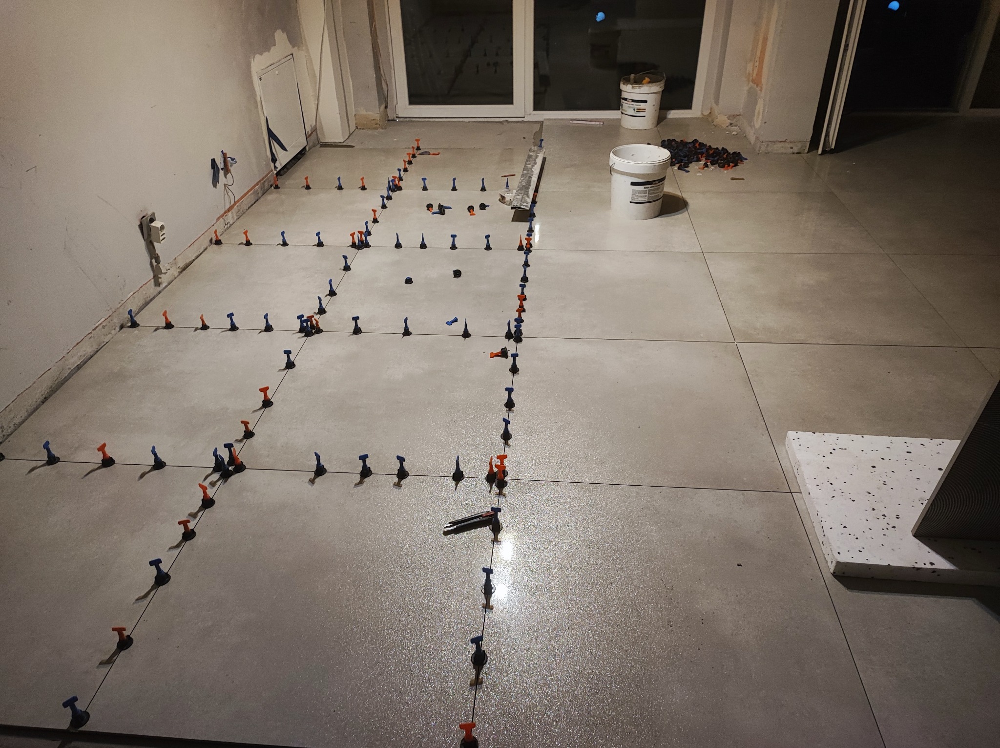 Układanie jasnoszarych płytek podłogowych z użyciem systemu poziomowania, widoczne krzyżyki dystansowe i wiaderka z zaprawą w tle.