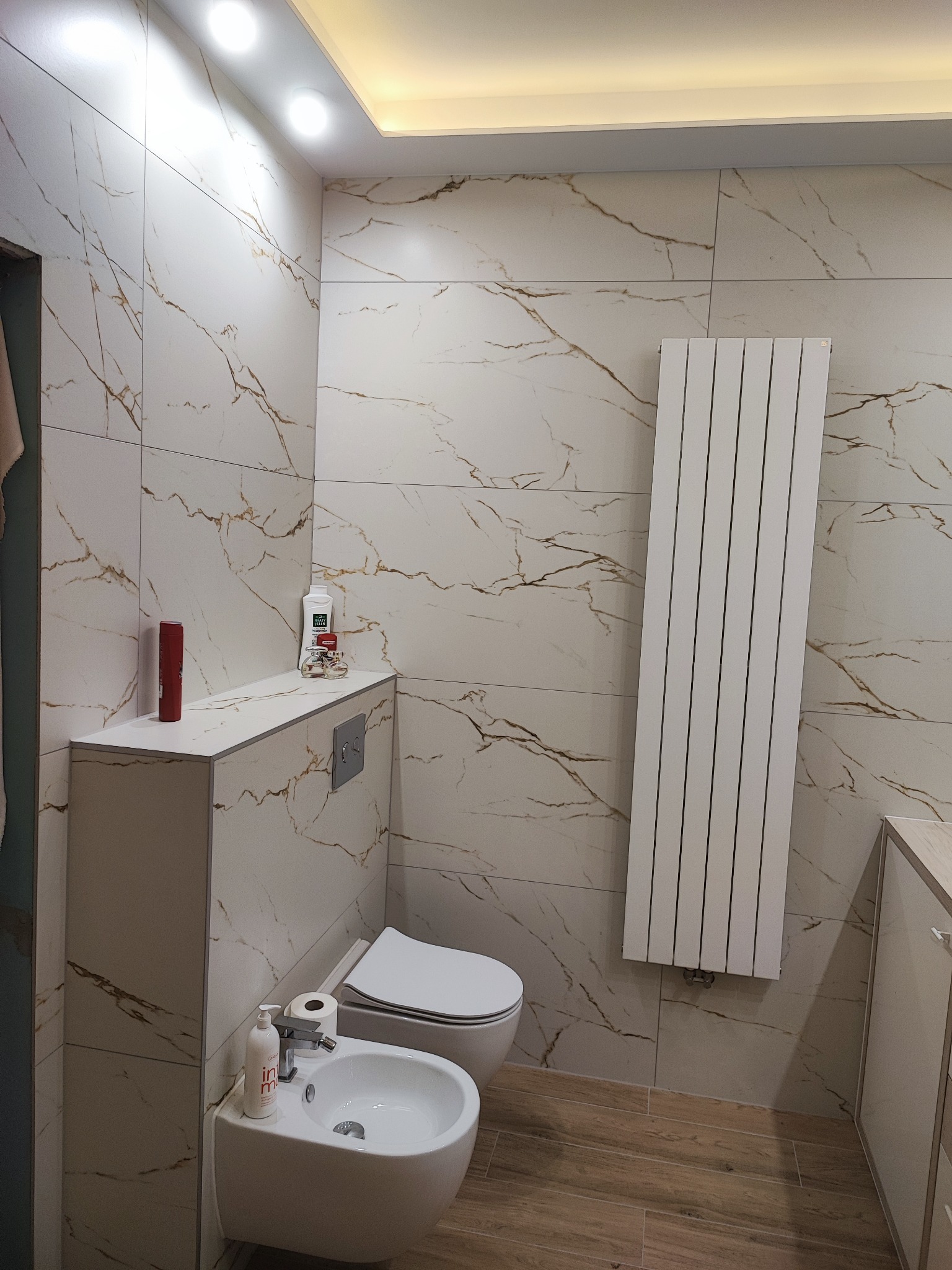 Wykończona łazienka w Nowogrodźcu z marmurowymi płytkami na ścianach, podwieszaną toaletą, bidetem i grzejnikiem panelowym.