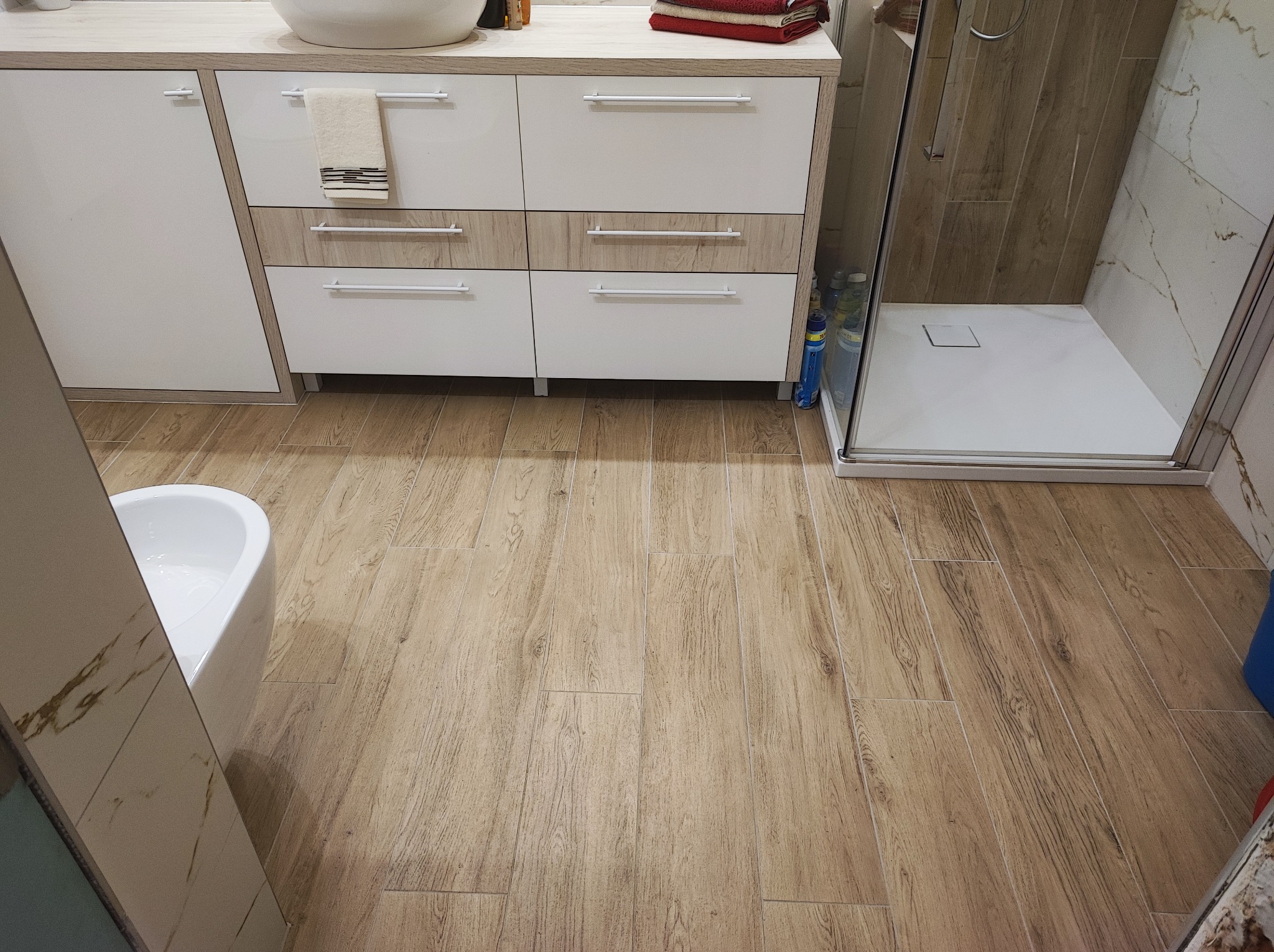 Nowoczesna łazienka z drewnopodobną podłogą, białą szafką pod umywalką z drewnianymi akcentami, kabiną prysznicową walk-in i częściowo widocznym bidetem.