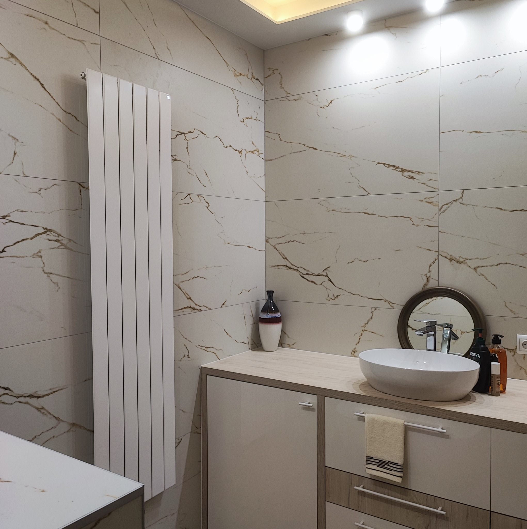 Nowoczesna łazienka z białymi płytkami imitującymi marmur, białym grzejnikiem panelowym, umywalką nablatową na szafce z jasnego drewna oraz okrągłym lustrem.