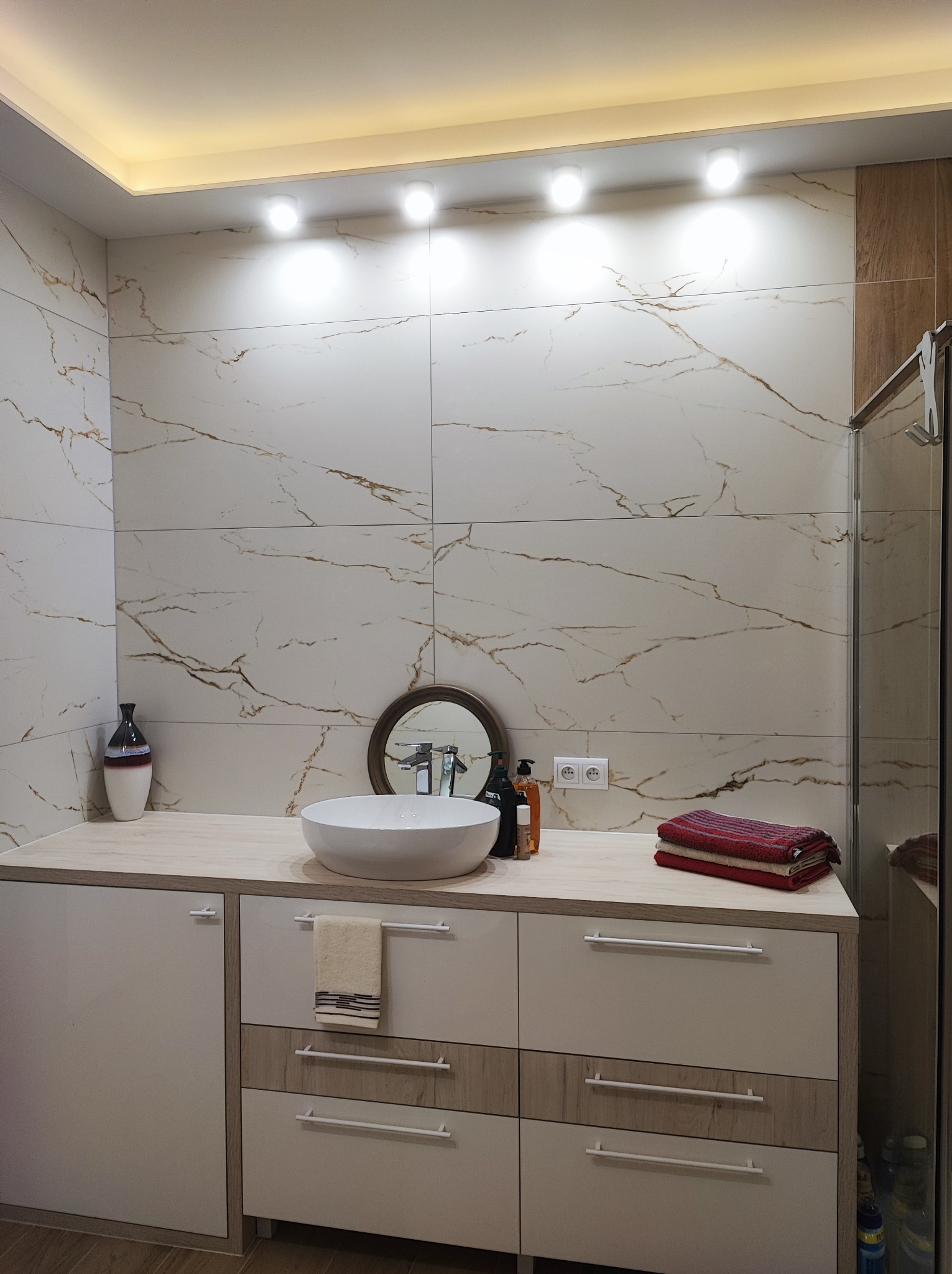 Nowoczesna łazienka z białymi płytkami imitującymi marmur z brązowymi żyłami, okrągła umywalka nablatowa, szafka pod umywalką z białymi frontami i drewnianymi akcentami, oświetlenie punktowe...