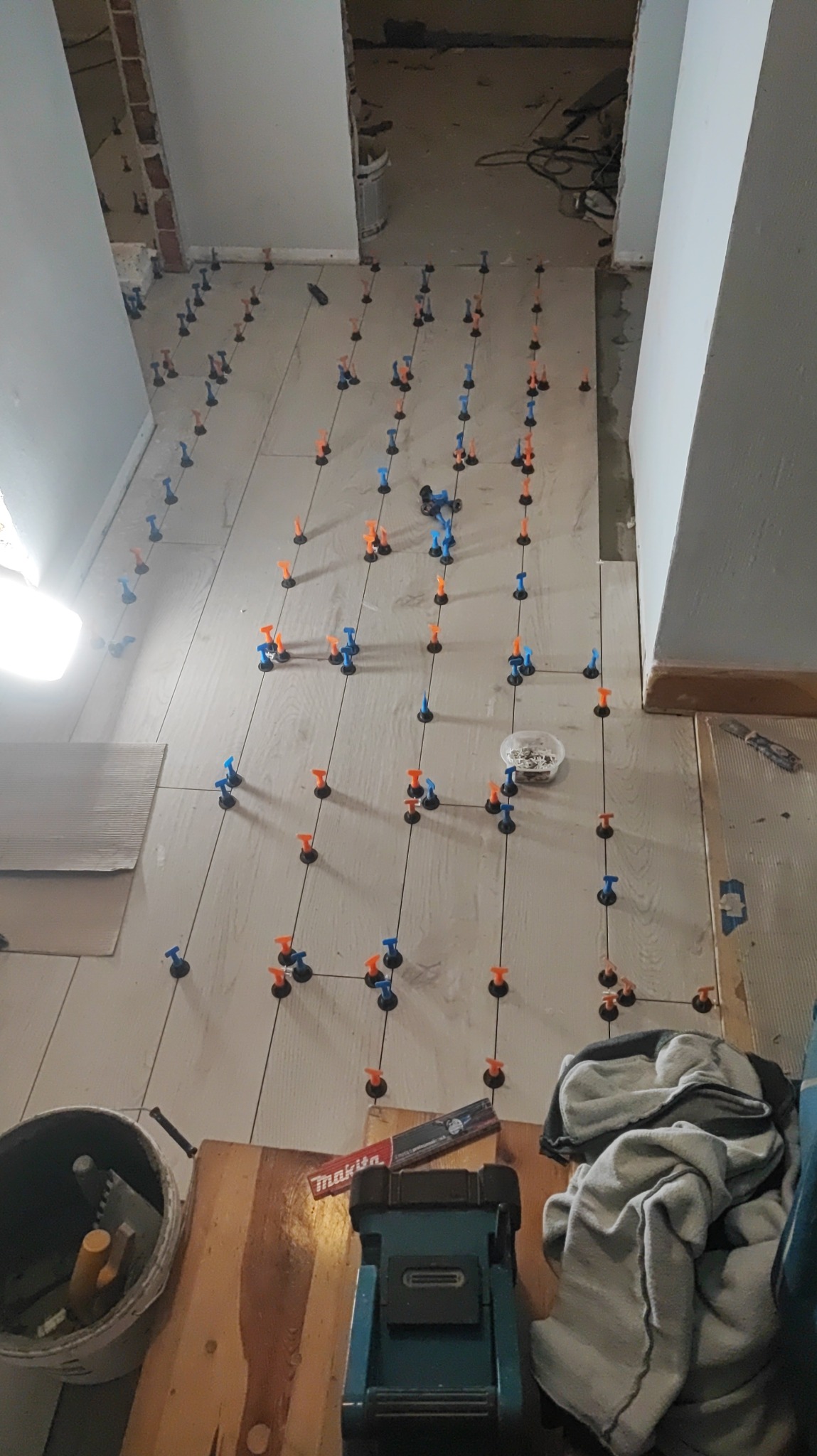 Układanie jasnych płytek podłogowych z użyciem systemu poziomowania płytek (klipsy i kliny) w trakcie remontu. Widoczny kubeł z narzędziami, poziomica i niebieska maszyna do cięcia płytek.