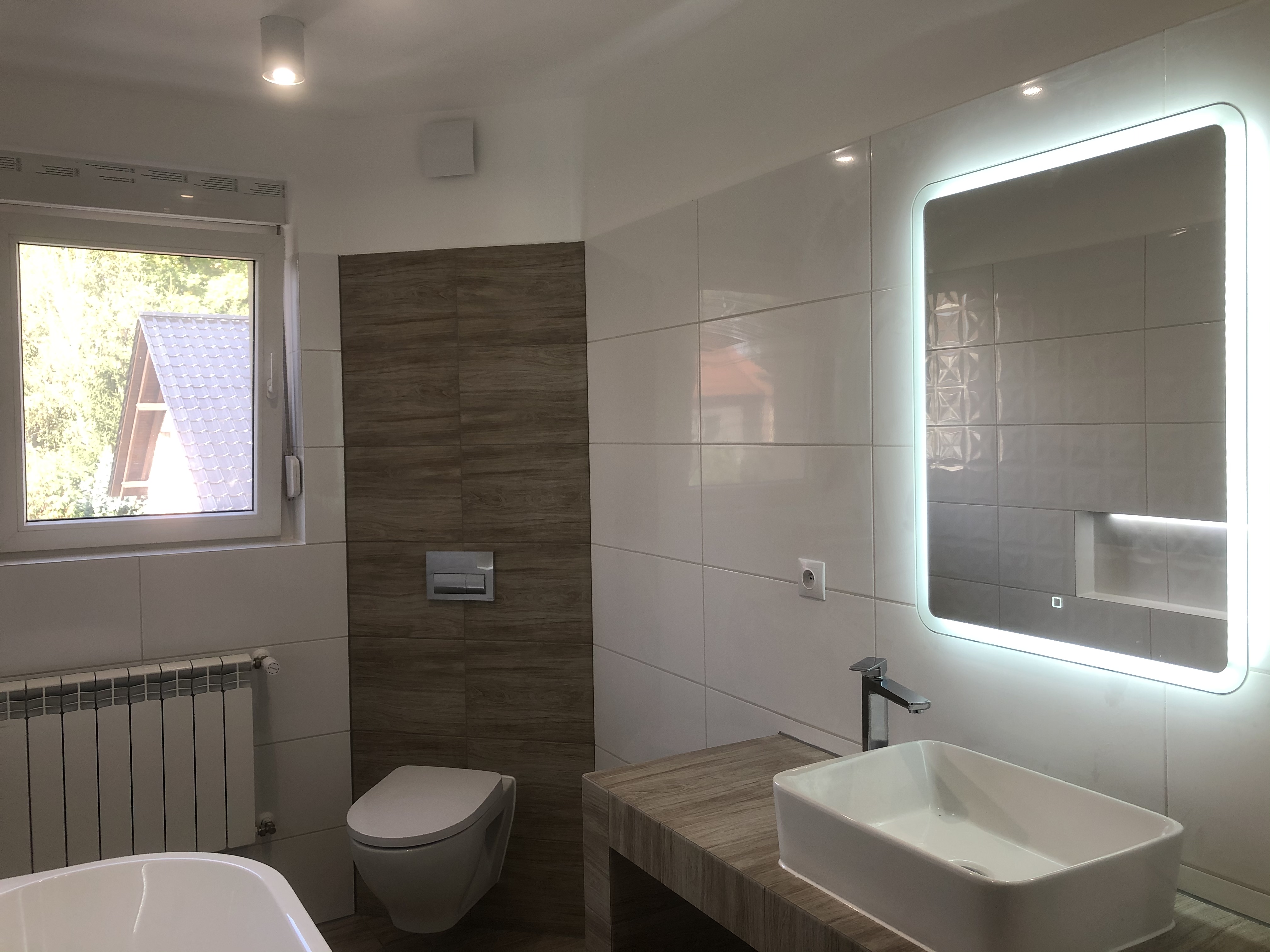 Nowoczesna łazienka z białymi i drewnopodobnymi płytkami, podświetlanym lustrem, wiszącą toaletą i widokiem z okna na dach domu.