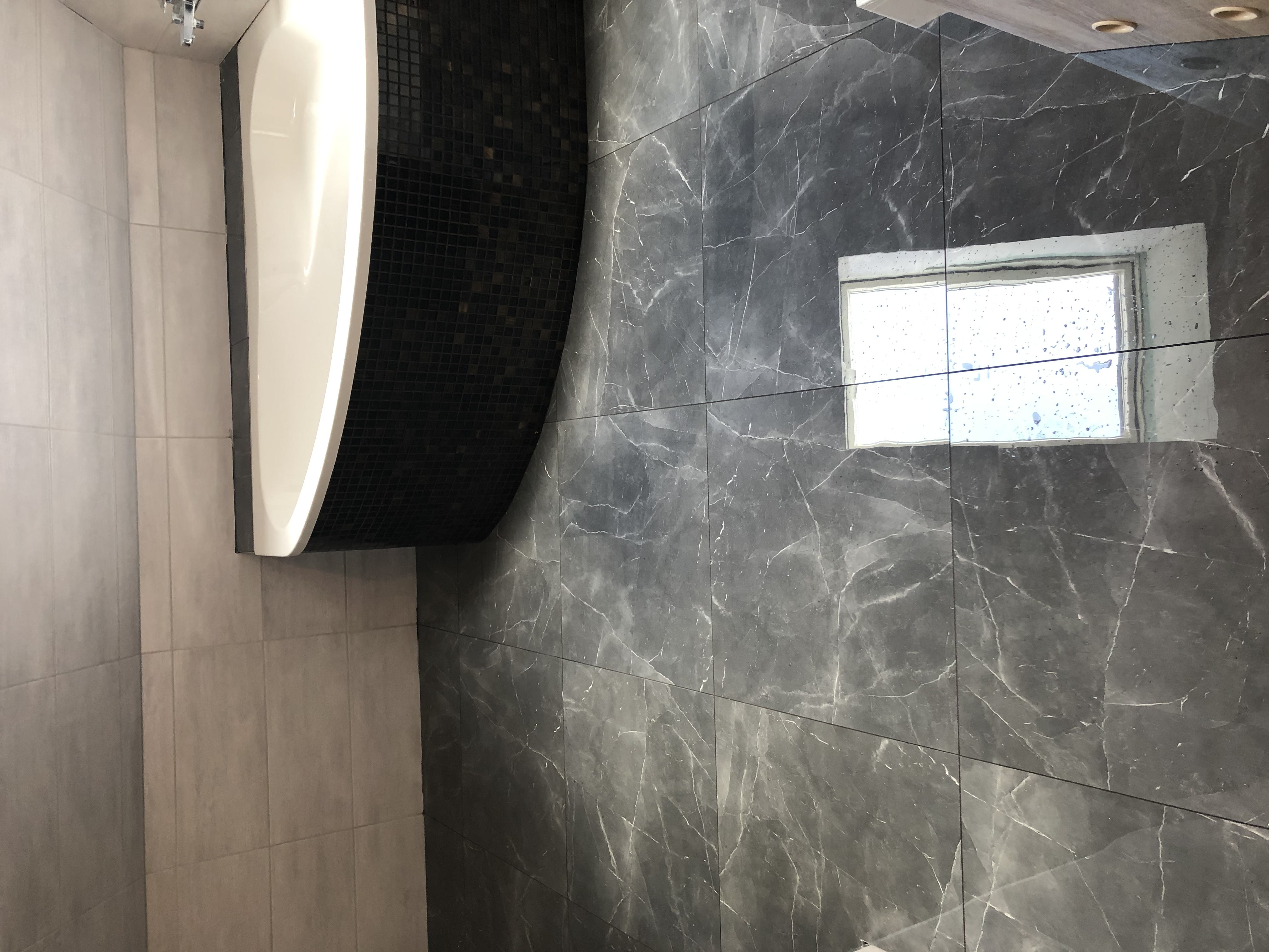 Nowoczesna łazienka z wanną, wykończona płytkami imitującymi marmur i mozaiką, z małym oknem z widokiem na zewnątrz.