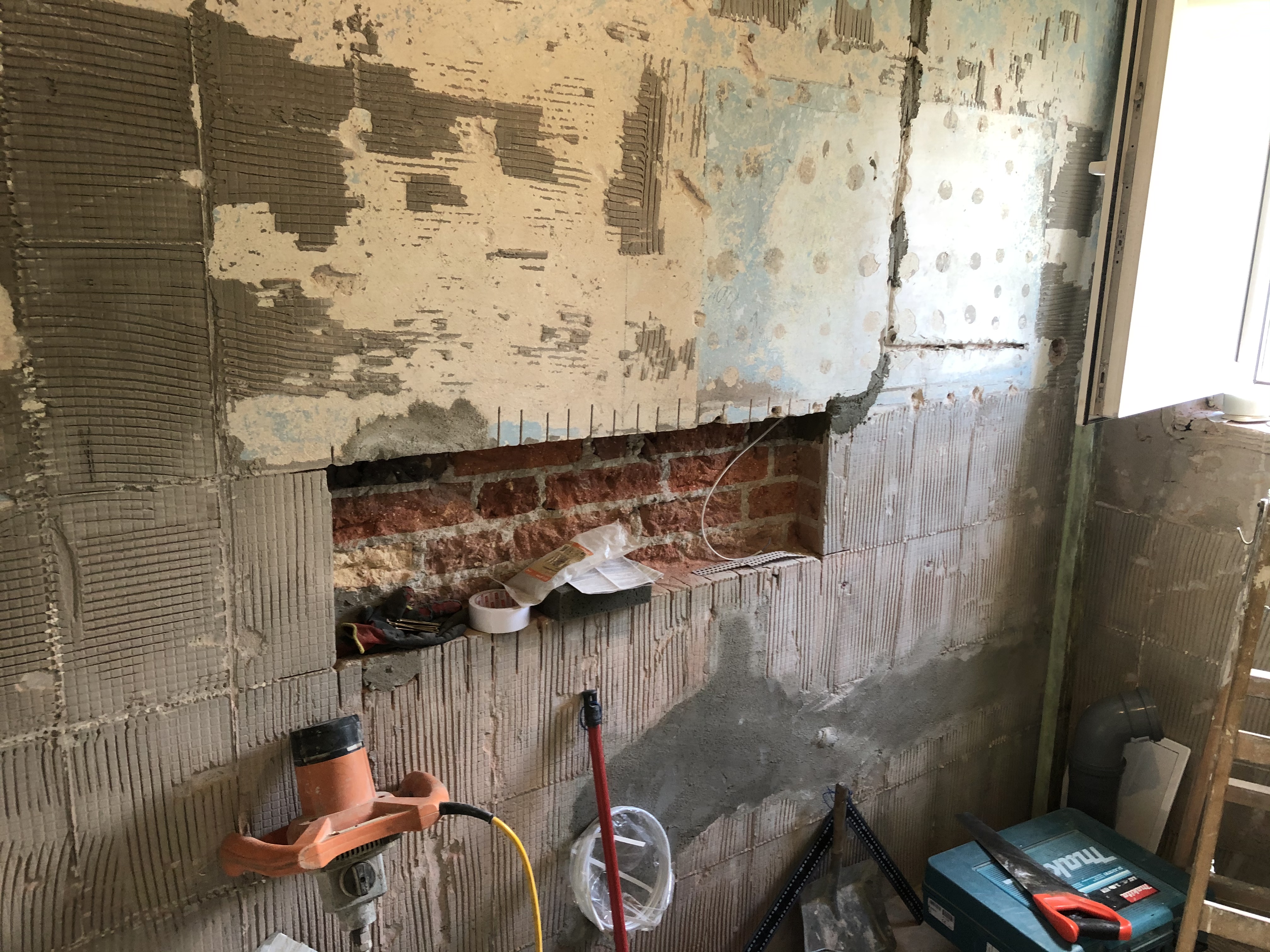 Przygotowanie ściany pod płytki w łazience: ściana z resztkami starej zaprawy i odkrytą czerwoną cegłą, widoczny mikser do zaprawy, narzędzia glazurnicze i poziomica laserowa.