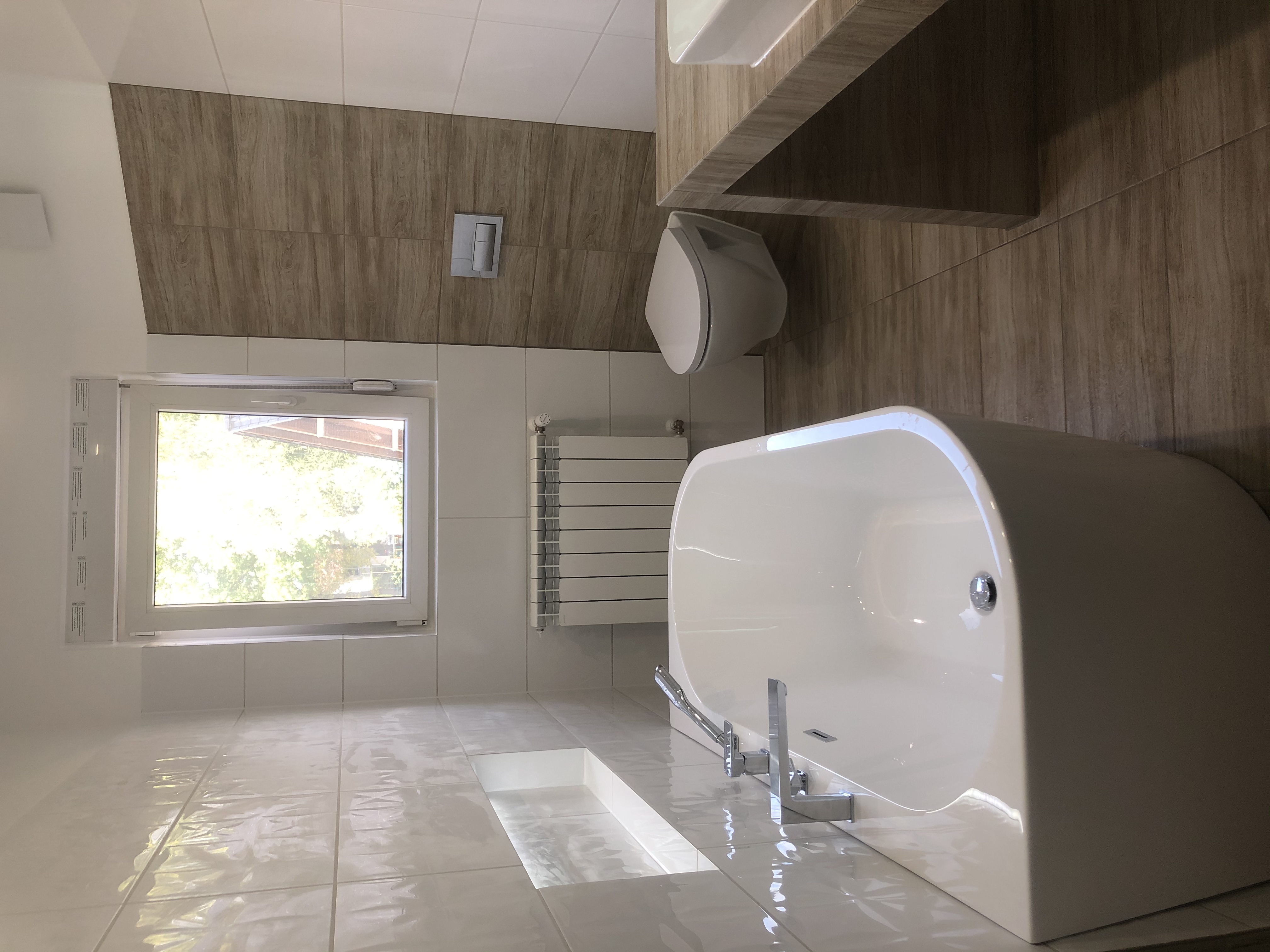 Nowoczesna łazienka z wolnostojącą wanną, podwieszaną toaletą, grzejnikiem panelowym i oknem. Ściany wyłożone płytkami w odcieniach bieli i drewna.