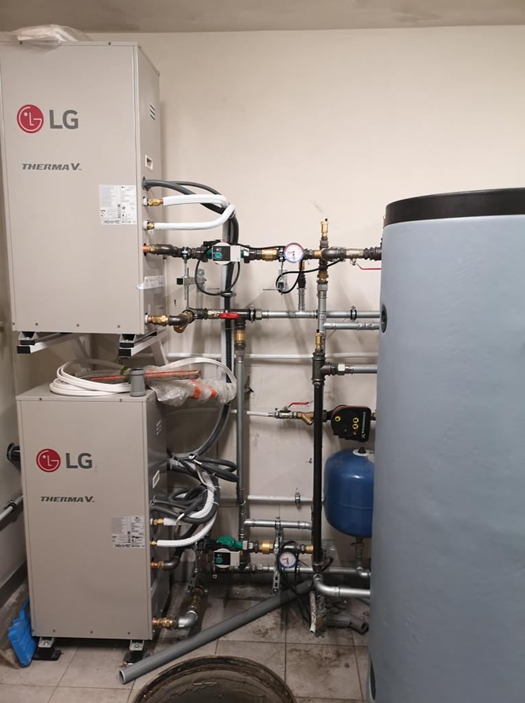 Dwa moduły pompy ciepła LG Therma V połączone rurami z instalacją grzewczą, w tle szary zasobnik ciepłej wody i niebieskie naczynie przeponowe.