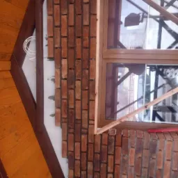 Murowana ściana z cegły klinkierowej w trakcie budowy, widoczne okno z drewnianą ramą, fragment drewnianej konstrukcji dachu i wystające przewody elektryczne.