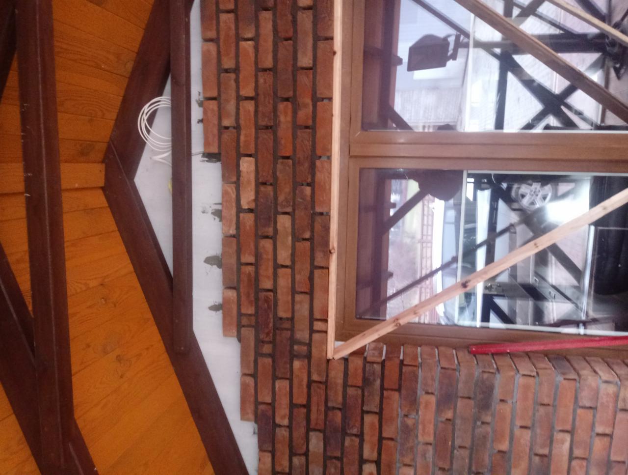 Murowana ściana z cegły klinkierowej w trakcie budowy, widoczne okno z drewnianą ramą, fragment drewnianej konstrukcji dachu i wystające przewody elektryczne.