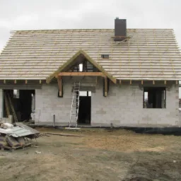 Niedokończony dom z bloczków, z drewnianą konstrukcją dachu i kominem, widoczna drabina oparta o wejście, materiały budowlane na palecie przed budynkiem, pochmurne niebo w tle.