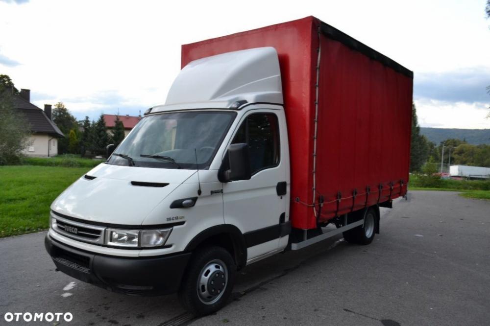 Biały samochód dostawczy Iveco z czerwoną plandeką na parkingu, widok z boku na tle domów i wzgórz.