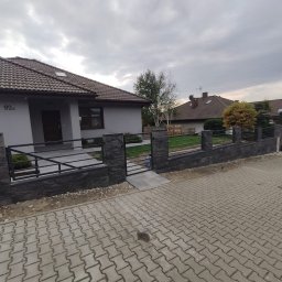Serwisbruk.pl - Nowoczesne ogrodzenie z czarnych, kamiennych elementów i metalowych przęseł, otaczające dom z szarą elewacją i brązowym dachem. Kostka brukowa na podjeździe.