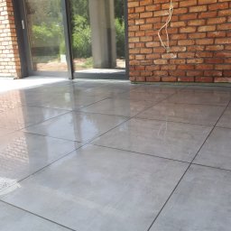 Serwisbruk.pl - Jasnoszara, błyszcząca posadzka z płytek na tarasie, przylegająca do ceglanej ściany i szklanych drzwi przesuwnych. Widoczne odbicia światła na powierzchni płytek.