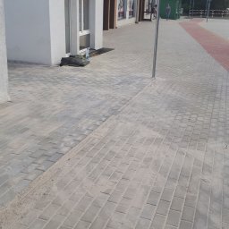 Serwisbruk.pl - Świeżo ułożona, szara kostka brukowa przed budynkiem z widocznymi elementami infrastruktury miejskiej, takimi jak chodnik z kostki czerwonej i ścieżka rowerowa.