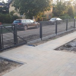 Serwisbruk.pl - Nowoczesne ogrodzenie panelowe z poziomych profili, brama wjazdowa, chodnik z kostki brukowej, w tle dom i samochody. Widoczne kosze na śmieci.
