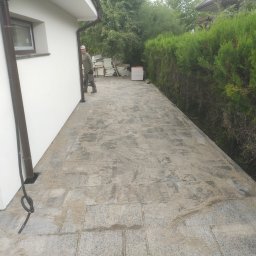 Serwisbruk.pl - Ułożona kostka brukowa wzdłuż białego budynku, widoczny fragment osoby i materiały budowlane w tle, żywopłot. Kostka zasypana piaskiem.