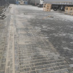 Serwisbruk.pl - Częściowo ułożona, szara kostka brukowa na parkingu dla tirów. Widoczne palety, narzędzia i pracownicy w tle. Część kostki zasypana piaskiem.