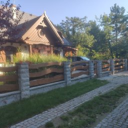 Serwisbruk.pl - Rustykalne ogrodzenie z kamiennymi słupkami i drewnianymi przęsłami, otaczające posesję z domem w stylu dworkowym. Kostka brukowa na podjeździe.