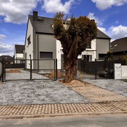 Serwisbruk.pl - Nowoczesny dom z szarą elewacją i brukowanym podjazdem z metalową bramą. Centralnie umieszczone drzewo dodaje naturalnego charakteru.