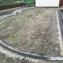 Serwisbruk.pl - Etap prac: obrzeża z ciemnej kostki ułożone wzdłuż ścieżki i rabaty, przygotowanie terenu pod dalsze prace brukarskie. Widoczny pracownik w tle.