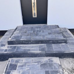 Serwisbruk.pl - Schody wejściowe z szarej kostki brukowej prowadzące do ciemnych drzwi wejściowych w białym budynku. Widoczne ślady piasku na kostce.