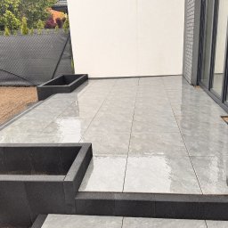 Serwisbruk.pl - Nowoczesny taras z szarych, kwadratowych płyt, obrzeża z czarnych elementów. Widok na ścianę budynku i fragment ogrodu z czarnym ogrodzeniem.