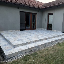Serwisbruk.pl - Wykończony taras z płytek ceramicznych z dwoma stopniami, przylegający do szarego budynku z brązowymi oknami i drzwiami. Widok na trawnik i fragment ogrodu.