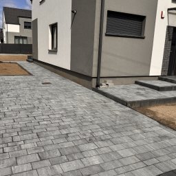 Serwisbruk.pl - Ułożona szara kostka brukowa prowadząca do nowoczesnego domu z szarymi elementami. Widoczny fragment ogrodu z przygotowanym podłożem pod trawnik.