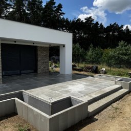 TarasyOgrody - Nowoczesny taras z szarych płyt, częściowo wykończony, z widoczną konstrukcją pod rabaty. W tle dom z ceglaną elewacją i las.