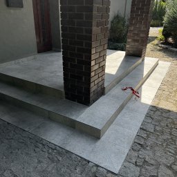 Serwisbruk.pl - Wykończone płytkami schody wejściowe z ceglanymi filarami, prowadzące do drzwi. Na zewnątrz widoczna kostka brukowa i zieleń. Ujęcie z dołu, podkreślające detale.