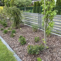 Serwisbruk.pl - Aranżacja ogrodu z szarą, poziomą, drewnianą palisadą, korą drzewną, niskimi krzewami i młodymi drzewkami. Obrzeże z kostki brukowej oddziela trawnik.