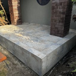 TarasyOgrody - Wykończony taras z płytkami ceramicznymi w odcieniach beżu i błękitu, z ceglanymi filarami. Widoczne elementy ogrodu i pojedyncza płytka na ziemi.
