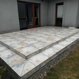 Serwisbruk.pl - Wykończony taras z dwoma stopniami, wyłożony wzorzystymi płytkami ceramicznymi w odcieniach beżu i błękitu. Widoczne drzwi tarasowe i okno. Na trawie stoją dwa pachołki.